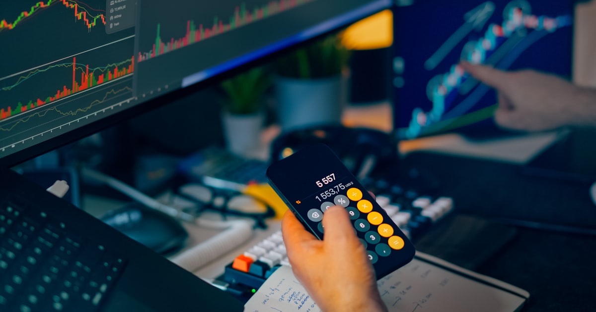 Trader analyzing stock market charts on computer screens with calculator.