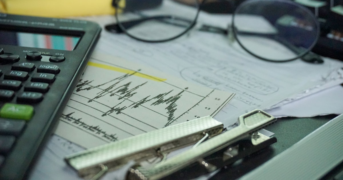 A desk with a calculator, papers, eyeglasses, and office supplies.