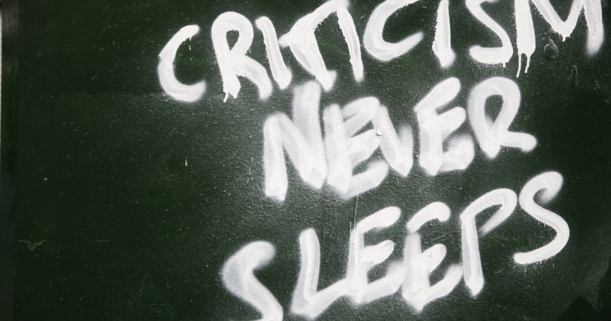 Graffiti on a green wall reads "criticism never sleeps"