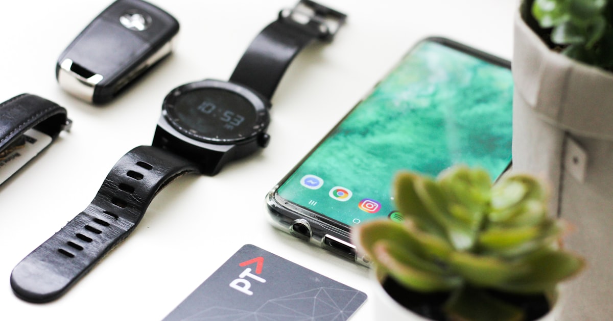 black Android smartphone near round black smartwatch, car fob, leather watch, and green succulent plant