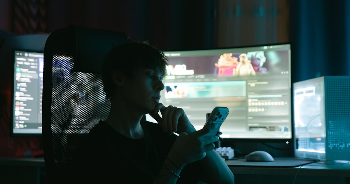 Person looking at phone in front of multiple computer monitors